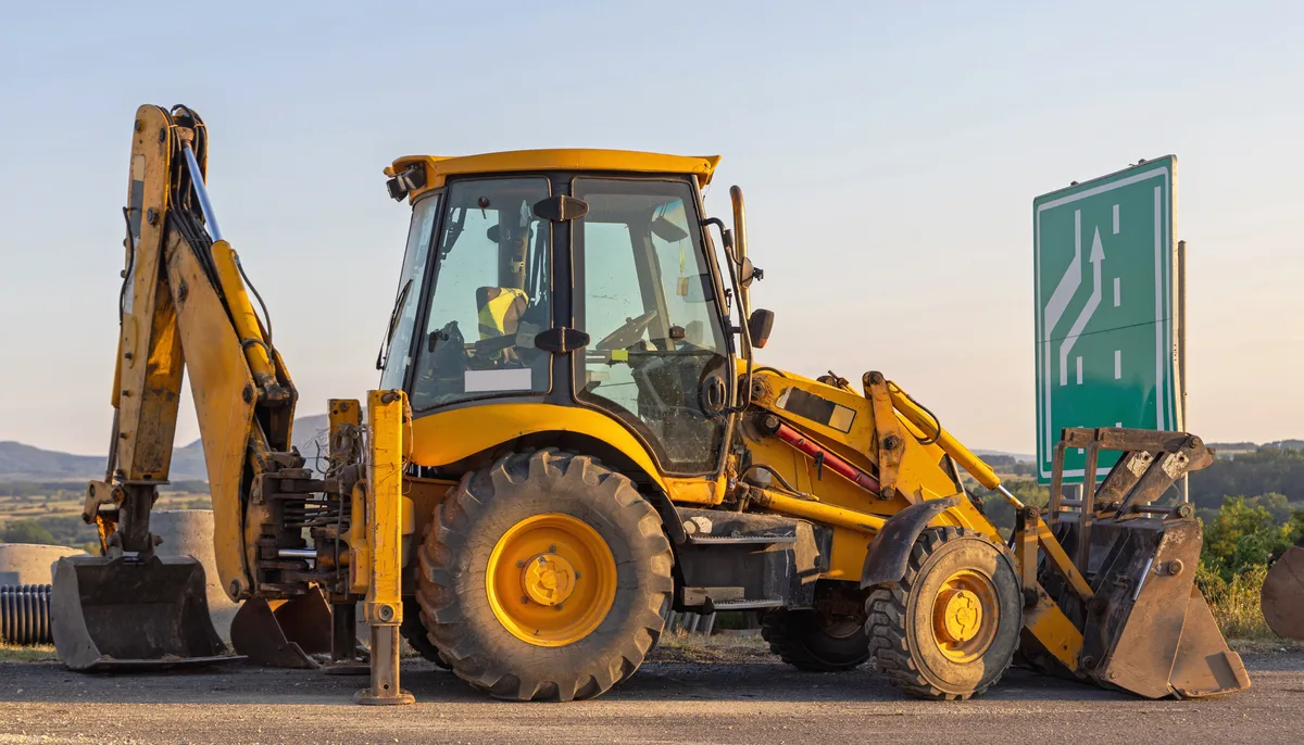 A backhoe loader with its rear boom raised, parked at a roadside under an evening sky — backhoe insurance in Indiana