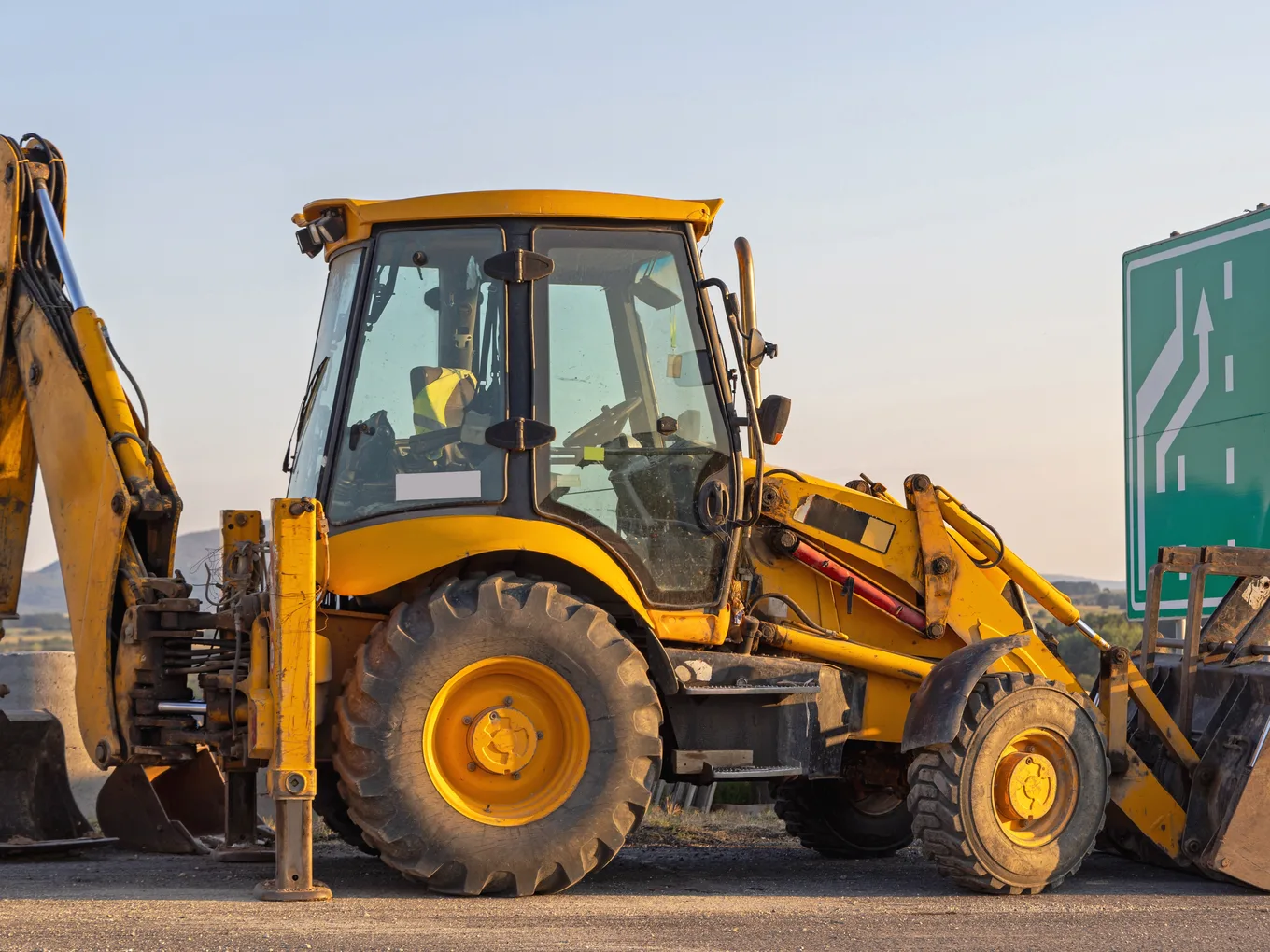 A backhoe loader with its rear boom raised, parked at a roadside under an evening sky — backhoe insurance in Delaware