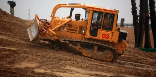 A crawler dozer with its blade lowered working reddish earth beside palms — dozer insurance in Oklahoma