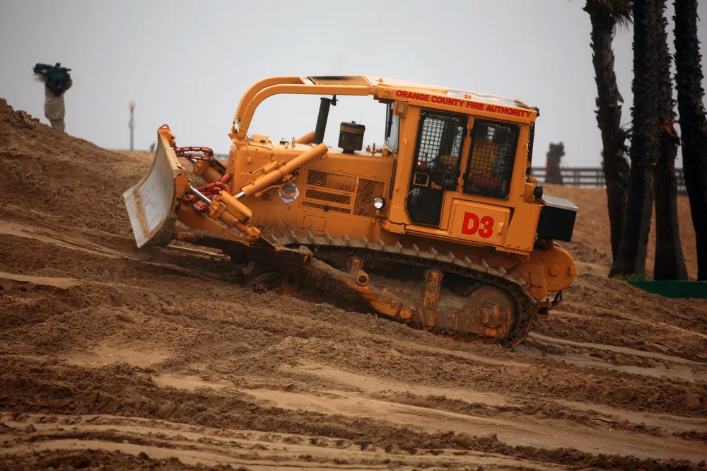 A crawler dozer with its blade lowered working reddish earth beside palms — dozer insurance in Missouri