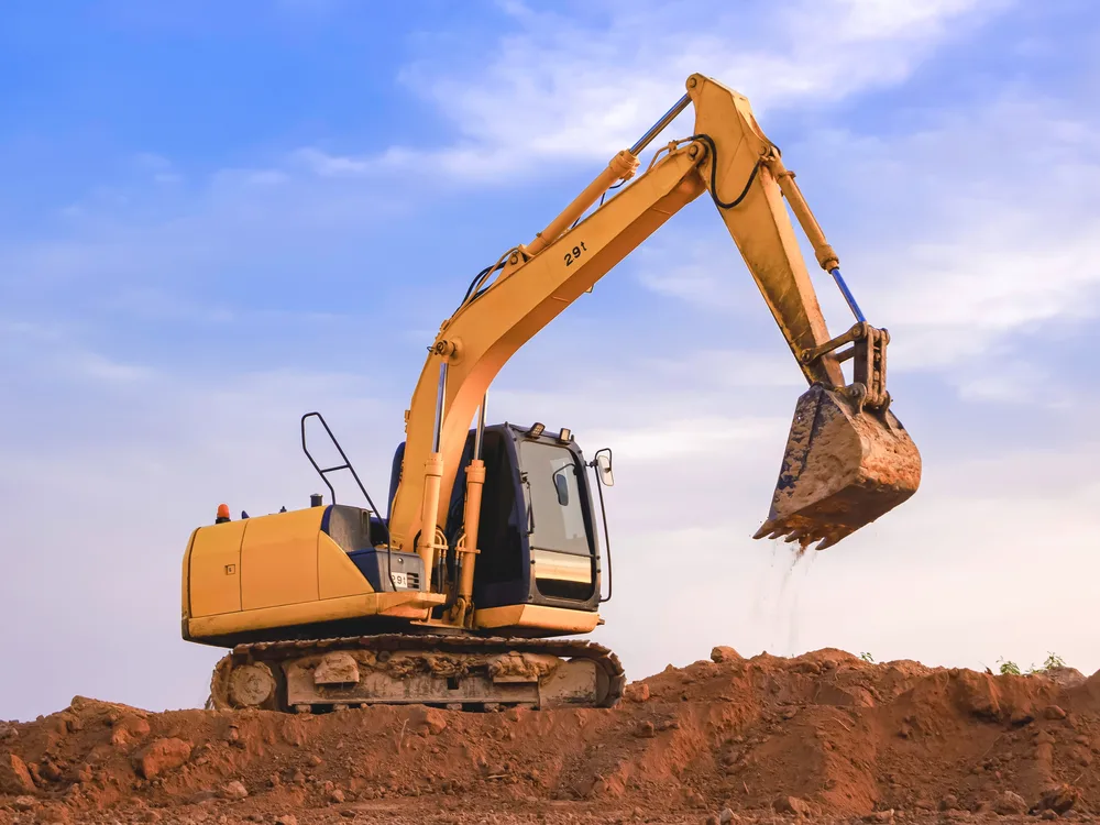 A tracked excavator with its arm raised on a mound of red earth against a blue sky — excavator insurance in Illinois