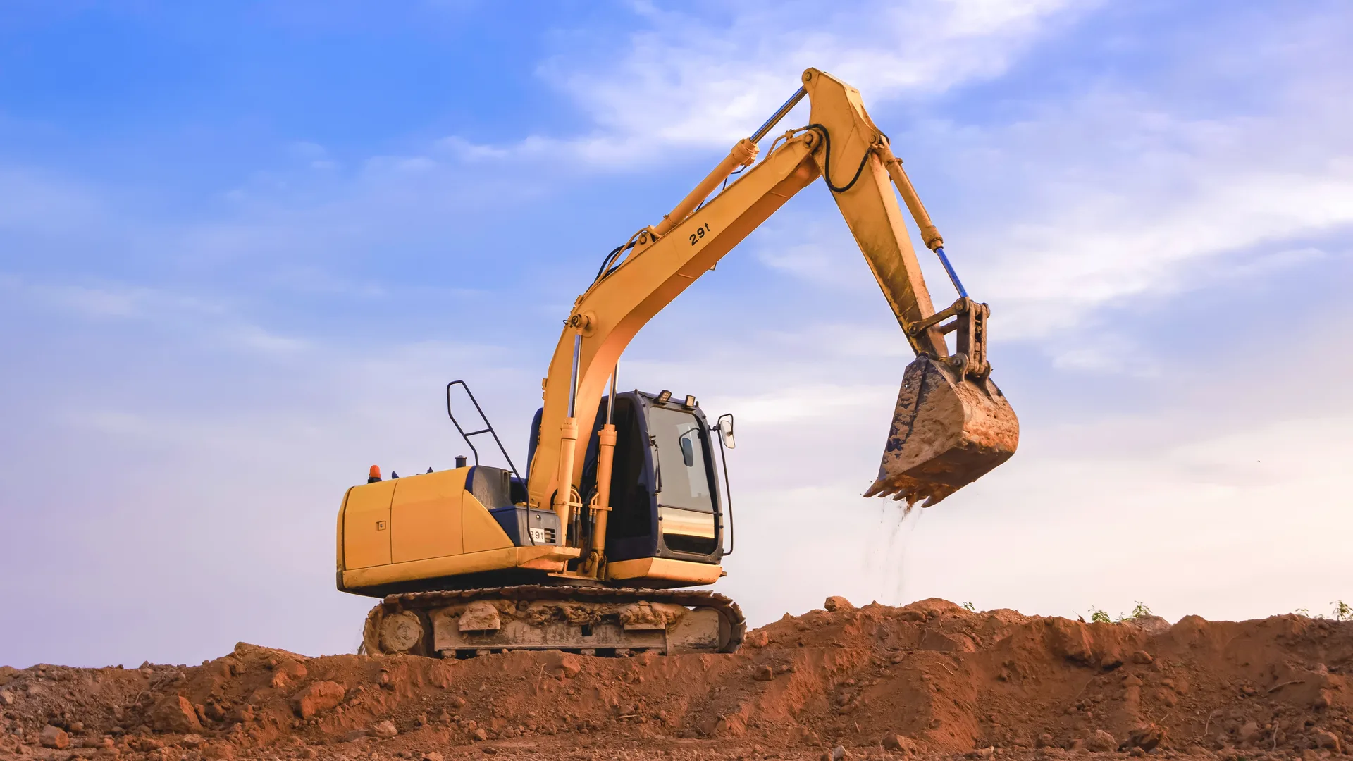 A tracked excavator with its arm raised on a mound of red earth against a blue sky — excavator insurance in Delaware