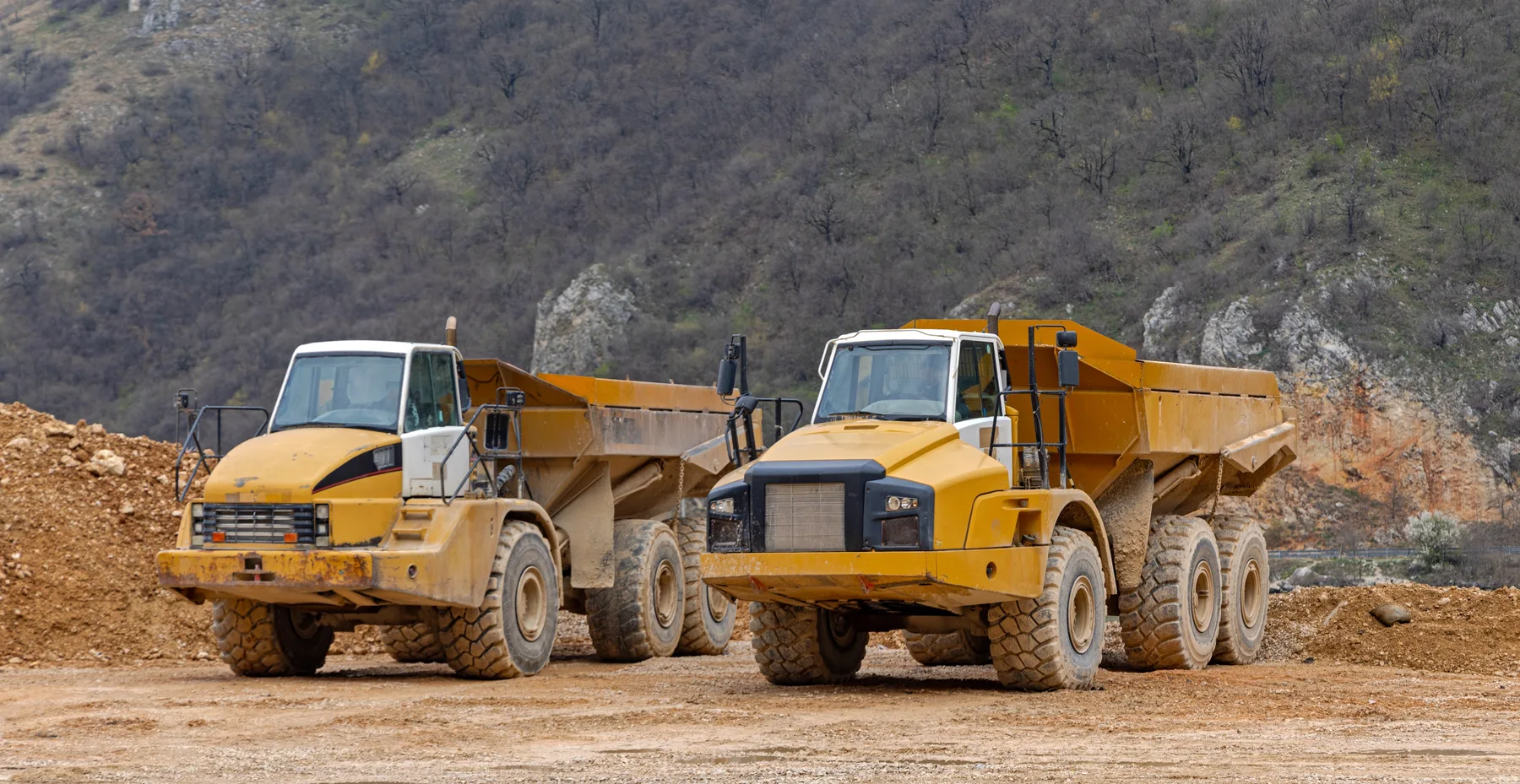 Articulated dump haulers parked on a dirt quarry floor below a rocky hillside