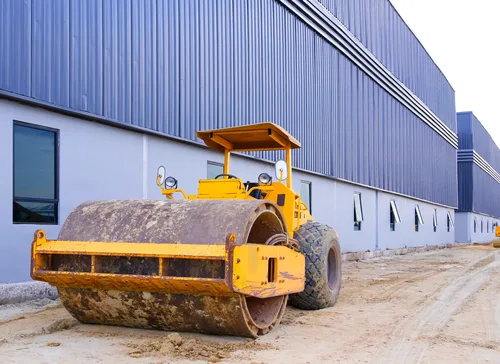 A drum roller compactor standing on a haul road beside a long clad warehouse wall