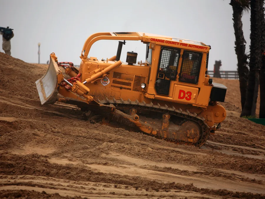 A crawler dozer with its blade lowered working reddish earth beside palms