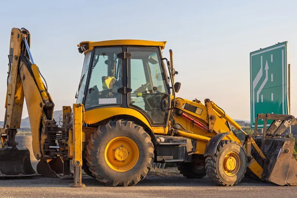 A backhoe loader with its rear boom raised, parked at a roadside under an evening sky