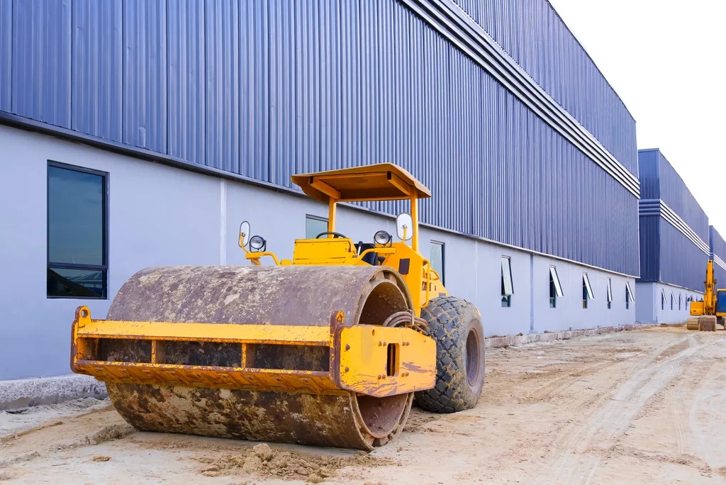 A drum roller compactor standing on a haul road beside a long clad warehouse wall