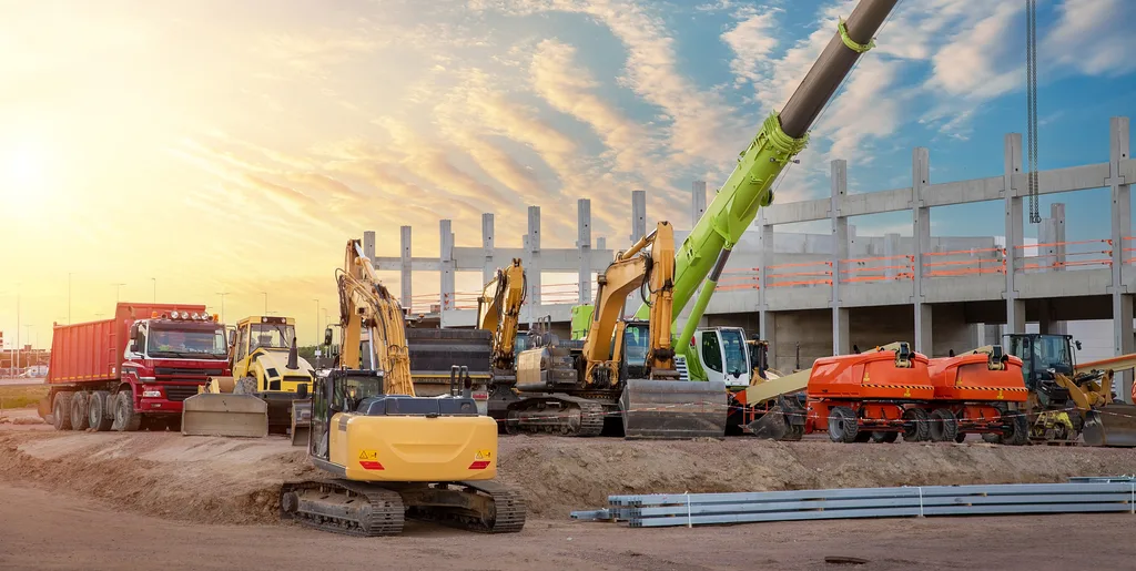 A line of excavators and haulers on a jobsite at sunset with a crane boom overhead