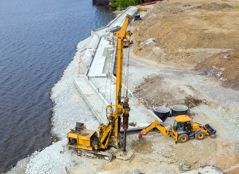 A piling rig on a stone embankment beside open water with a support machine alongside
