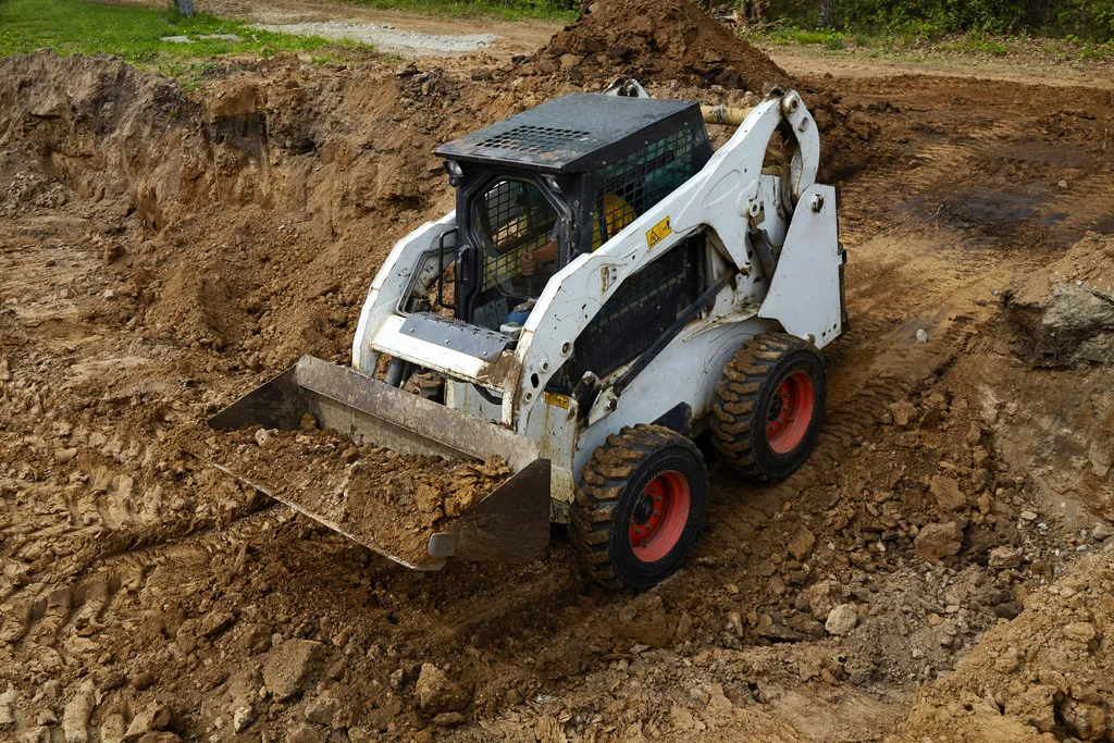 A skid steer loader with a raised bucket working a mound of dark earth