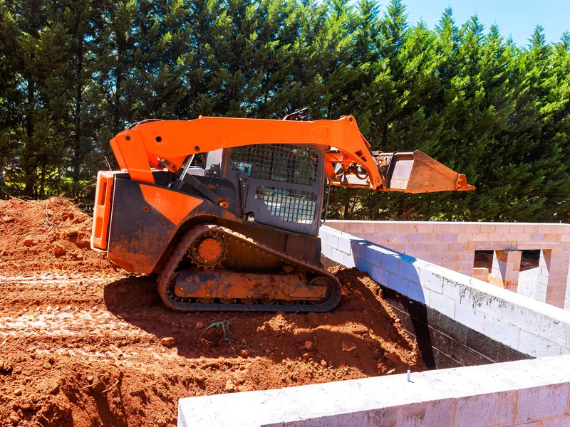 A compact track loader with a raised bucket beside a concrete foundation wall