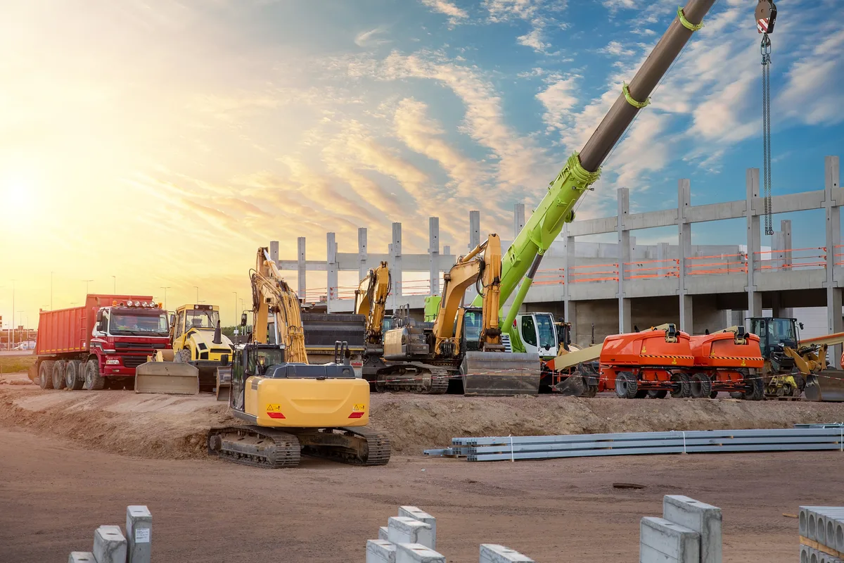 A line of excavators and haulers on a jobsite at sunset with a crane boom overhead
