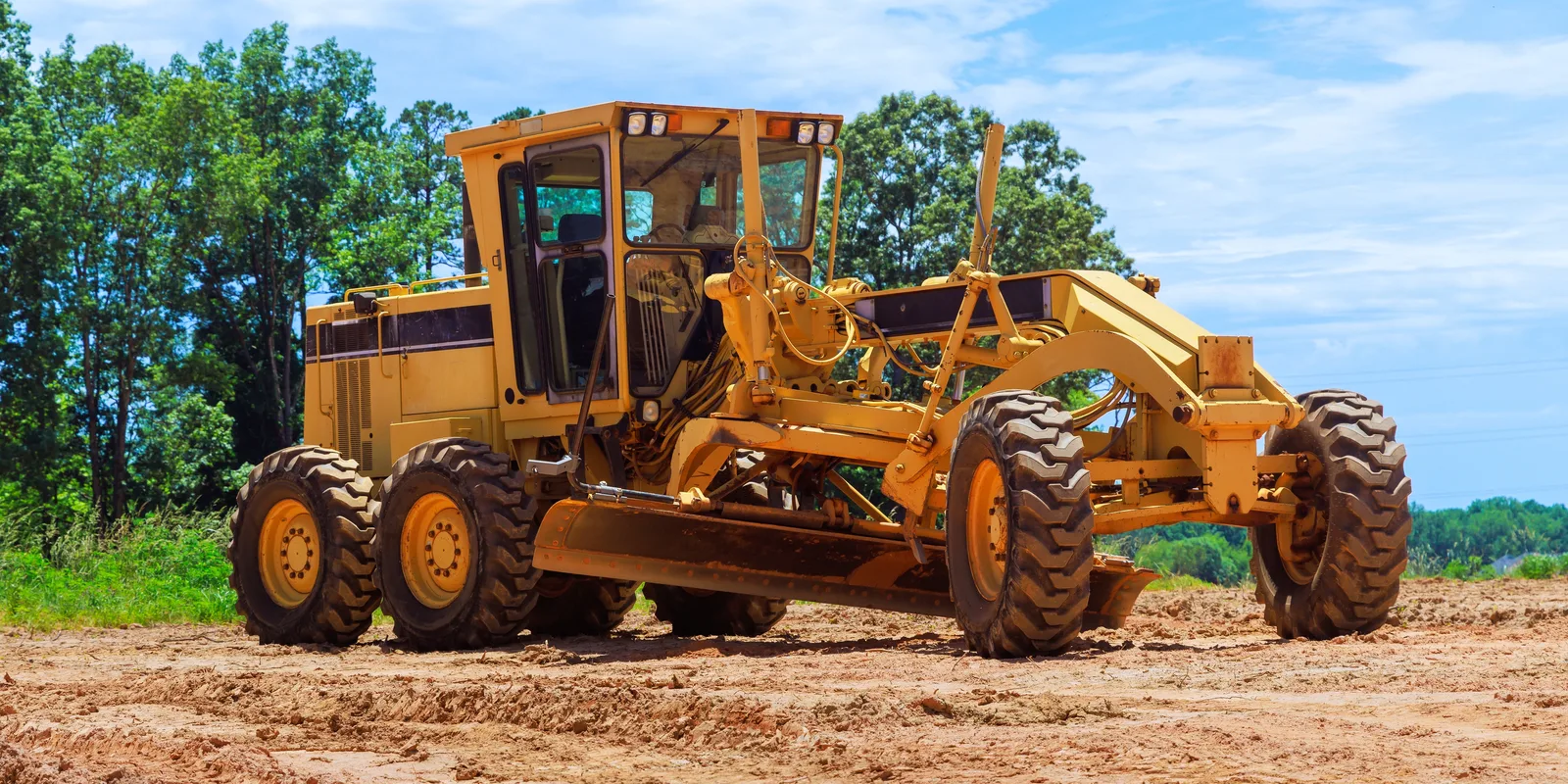 A motor grader standing on open dirt ground with trees behind under a clear sky — heavy equipment insurance in North Carolina
