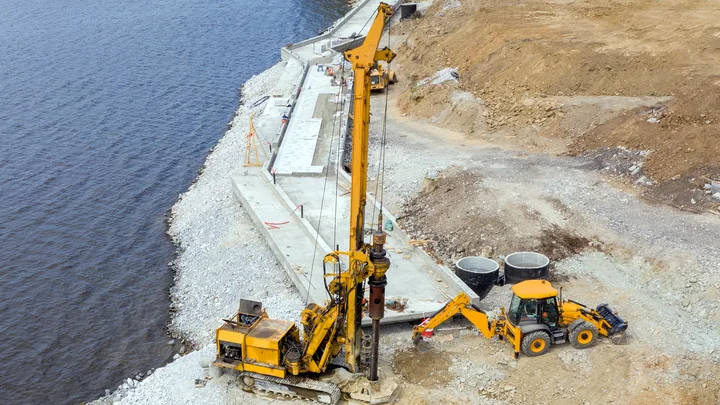 A piling rig on a stone embankment beside open water with a support machine alongside — heavy equipment insurance in Florida