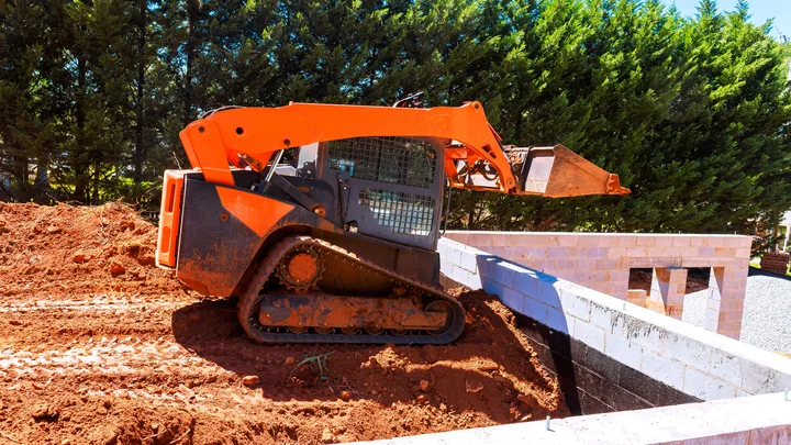 A compact track loader with a raised bucket beside a concrete foundation wall — heavy equipment insurance in California