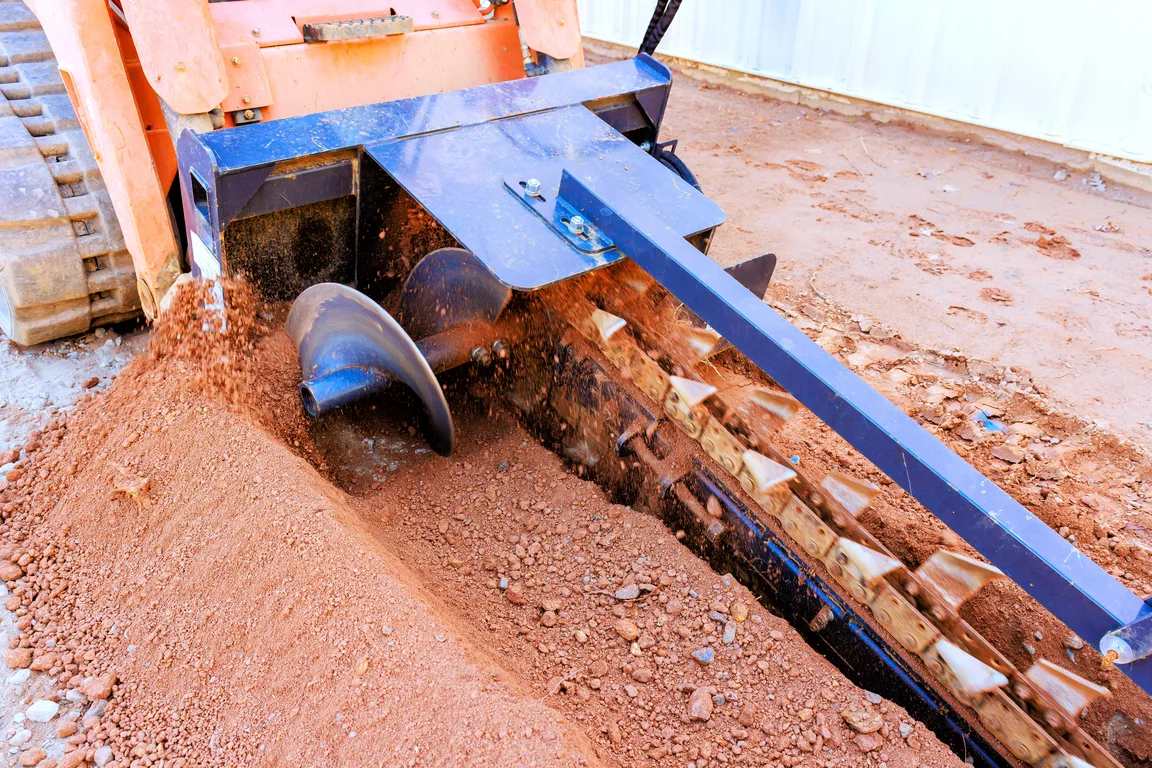 A trencher digging wheel cutting an open trench through red soil — heavy equipment insurance in Illinois