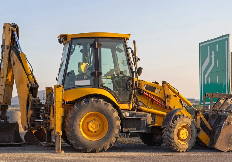 A backhoe loader with its rear boom raised, parked at a roadside under an evening sky