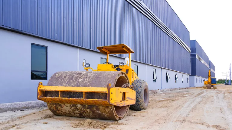 A drum roller compactor standing on a haul road beside a long clad warehouse wall