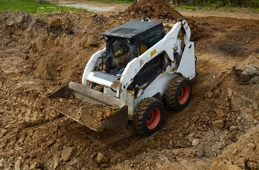 A skid steer loader with a raised bucket working a mound of dark earth — skid steer insurance in Ohio