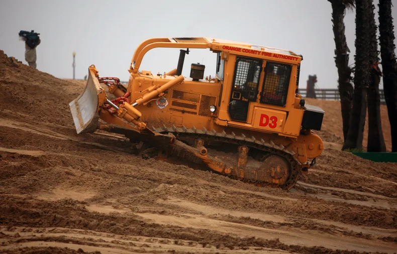 A crawler dozer with its blade lowered working reddish earth beside palms