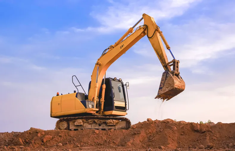 A tracked excavator with its arm raised on a mound of red earth against a blue sky