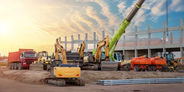 A line of excavators and haulers on a jobsite at sunset with a crane boom overhead