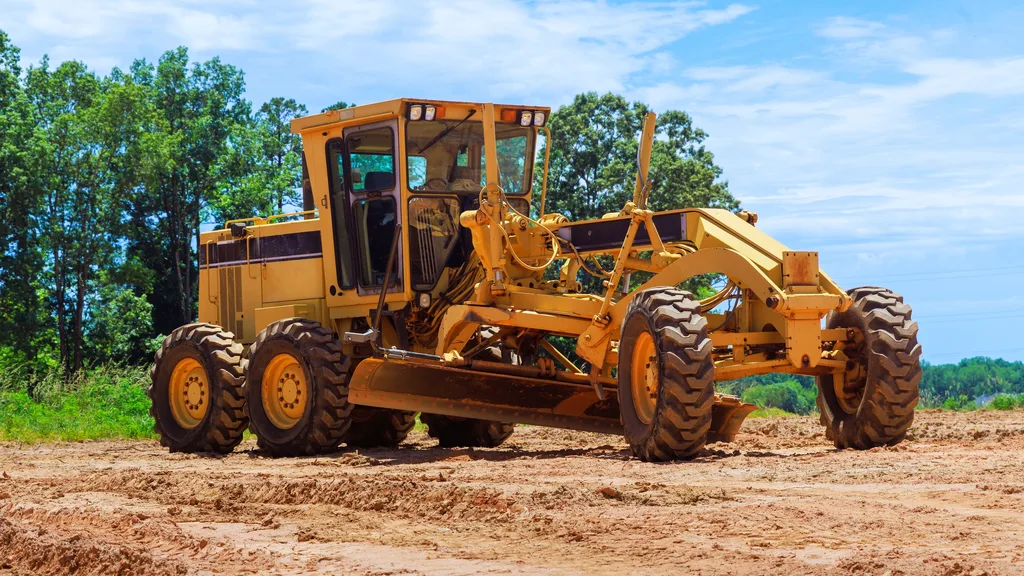 A motor grader standing on open dirt ground with trees behind under a clear sky