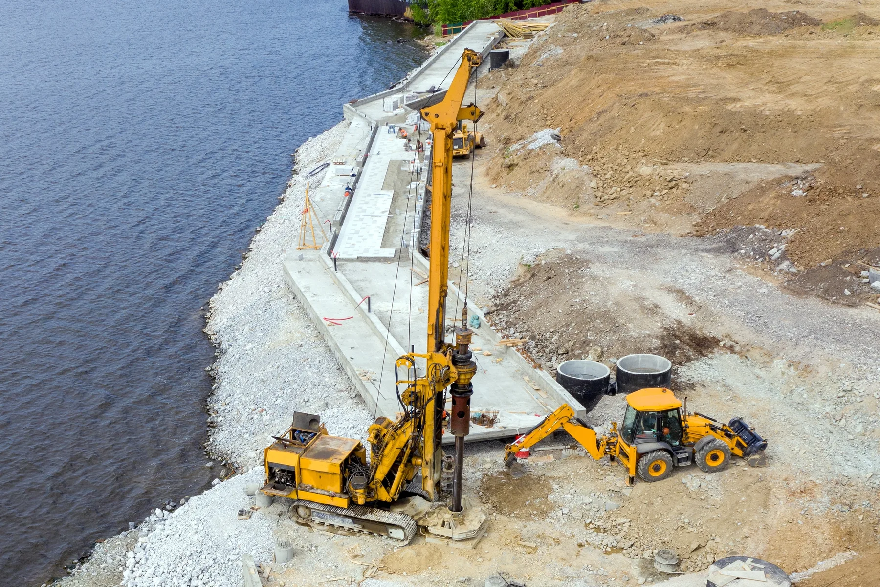 A piling rig on a stone embankment beside open water with a support machine alongside